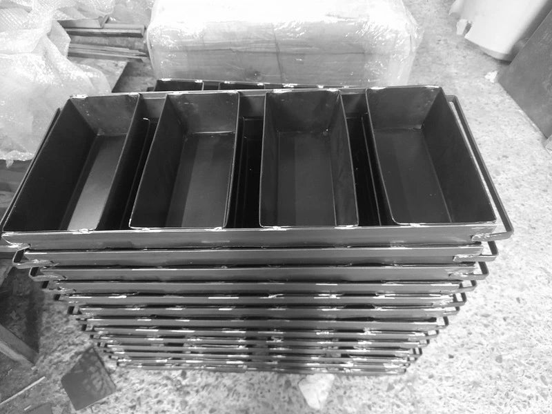 Stack of industrial 4-strap bread loaf molds in a workshop, made from heavy-duty black steel for commercial bakery use.